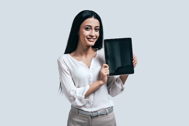 ¡Siempre ahí para ayudarme! Atractiva mujer joven en camisa blanca sosteniendo tableta digital y sonriendo mientras está de pie contra el fondo gris