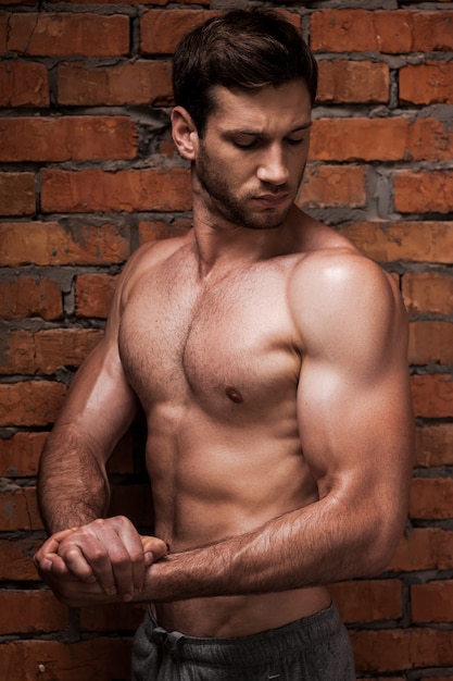 Siempre en buena forma. Hombre musculoso joven confiado posando mientras está de pie contra la pared de ladrillo