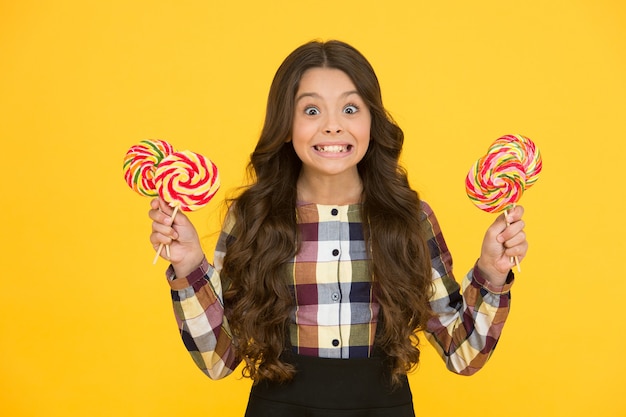 Siempre coma el postre primero. Feliz pequeño alumno mantenga postre de caramelo en palos. Sonrisa de niña con piruletas. Postre dulce. Merienda escolar. Su postre favorito en forma de remolino de arcoíris.