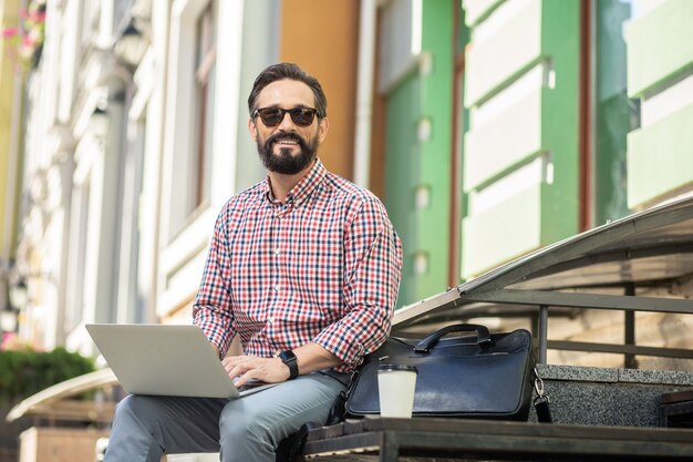 Siempre conectado. Hombre sonriente alegre sentado en la calle mientras usa una computadora portátil