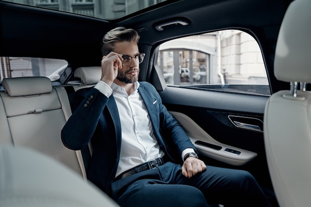 Siempre confiado. Apuesto joven en traje completo ajustando sus gafas mientras está sentado en el coche