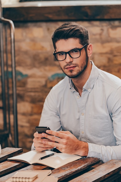 Siempre en contacto Apuesto joven pensativo sosteniendo un teléfono inteligente y mirando a la cámara mientras está sentado en su lugar de trabajo