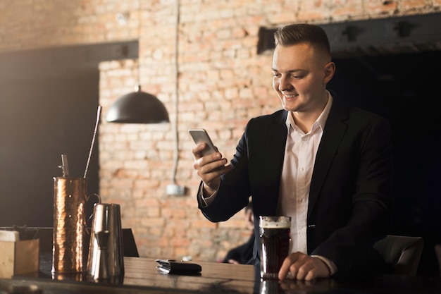 Siempre en contacto. Hombre alegre confiado sentado en la barra del bar, bebiendo cerveza y redes en el teléfono inteligente, espacio de copia