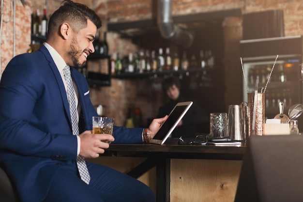 Siempre en contacto. Hombre de negocios alegre confiado que se sienta en la barra del bar, bebiendo whisky y trabajando en la tableta digital, espacio de la copia