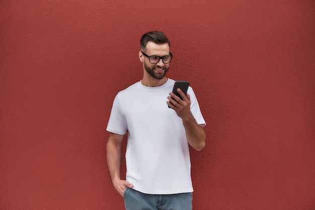 Siempre en contacto, un joven apuesto con ropa informal sosteniendo un teléfono móvil y sonriendo mientras