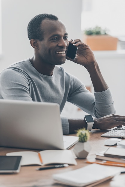 Siempre disponible. Apuesto joven africano hablando por teléfono inteligente y sonriendo mientras está sentado en su lugar de trabajo en la oficina moderna