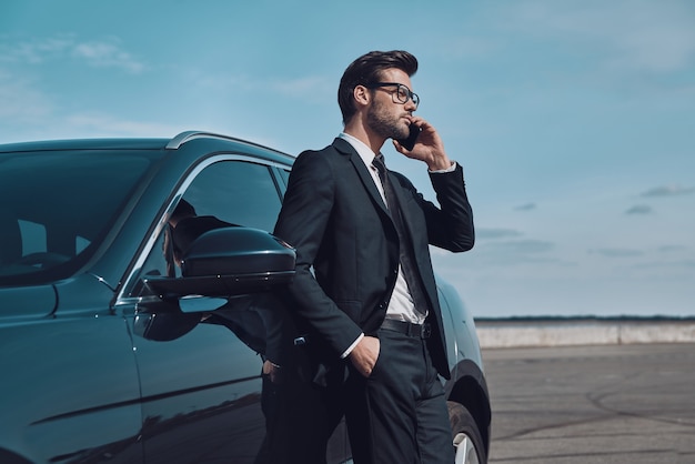 Siempre disponible. Apuesto joven empresario hablando por teléfono mientras está de pie cerca de su coche al aire libre