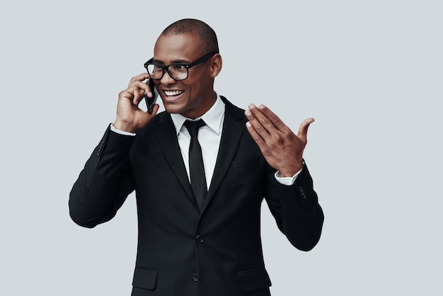 Siempre dispuesto a ayudar. Encantador joven africano en ropa formal hablando por teléfono inteligente y sonriendo mientras está de pie contra el fondo gris