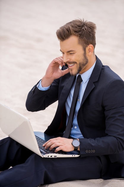 Siempre está en contacto. Alegre joven empresario trabajando en una computadora portátil y hablando por teléfono móvil mientras está sentado en la arena