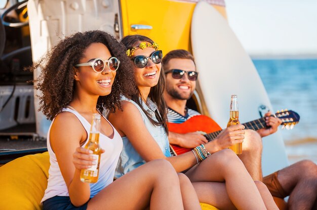 Siempre felices juntos. Tres jóvenes alegres bebiendo cerveza y tocando la guitarra mientras están sentados en la playa cerca de la furgoneta retro