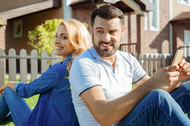 Siempre juntos. Amable pareja de mediana edad sentados espalda con espalda en el césped, volviendo la cabeza el uno al otro y sonriendo mientras el hombre sostiene un teléfono