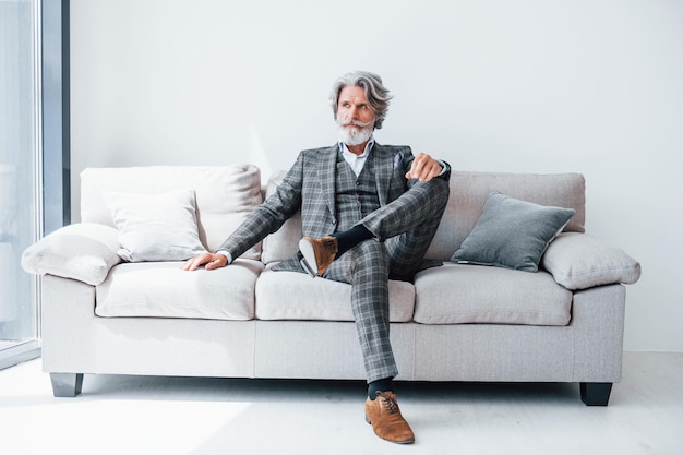 Se sienta en un cómodo sofá con ropa formal Hombre moderno elegante senior con pelo gris y barba en el interior