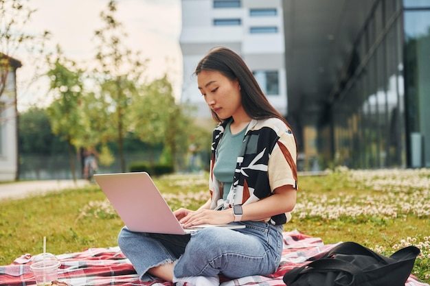 Se sienta con la computadora portátil La mujer asiática joven está al aire libre durante el día