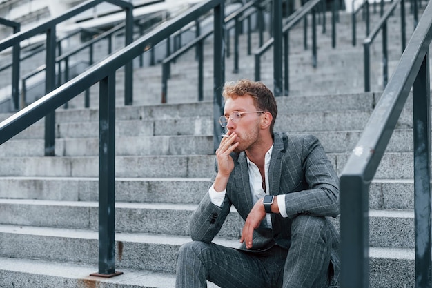 Se sienta y fuma cigarrillo Joven empresario exitoso en ropa formal gris está al aire libre en la ciudad