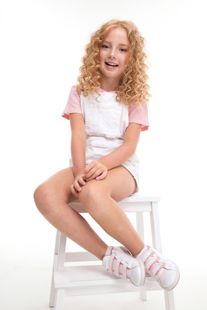 Se sienta una niña con el pelo rojo en una camisa, un mono blanco y zapatillas blancas.