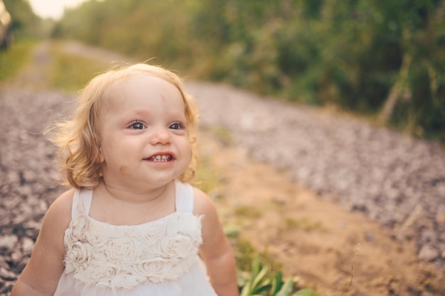 Se sienta el niño pequeño divertido lindo rubio niña con rizos en vestido blanco y con barro en la cara