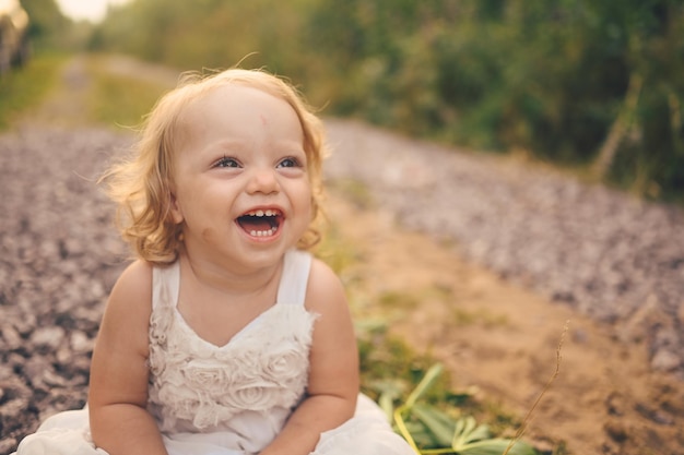 Se sienta el niño pequeño divertido lindo rubio niña con rizos en vestido blanco y con barro en la cara