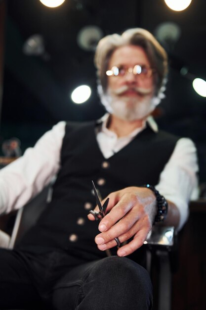 Se sienta en la silla en la barbería Elegante hombre senior moderno con cabello gris y barba está en el interior