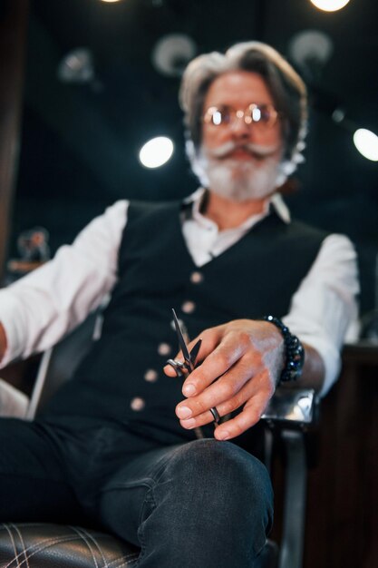 Se sienta en la silla en la barbería Elegante hombre senior moderno con cabello gris y barba está en el interior