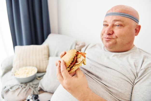 Se sienta en el sofá con palomitas de maíz y hamburguesas Un hombre gracioso con sobrepeso vestido con ropa informal está en el interior de su casa