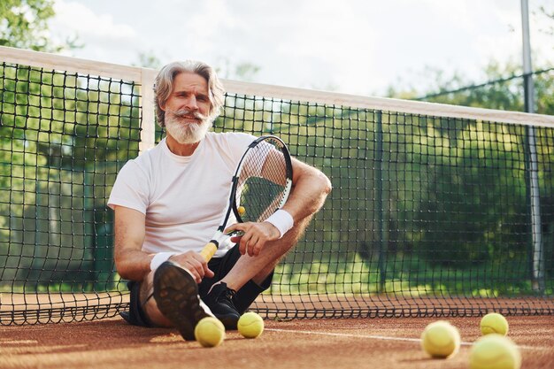 Se sienta en el suelo y toma un descanso Hombre moderno y elegante con raqueta al aire libre en la cancha de tenis durante el día