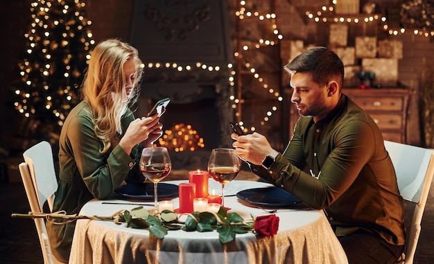 Se sienta y usa el teléfono Encantadora pareja joven tiene una cena romántica en el interior juntos