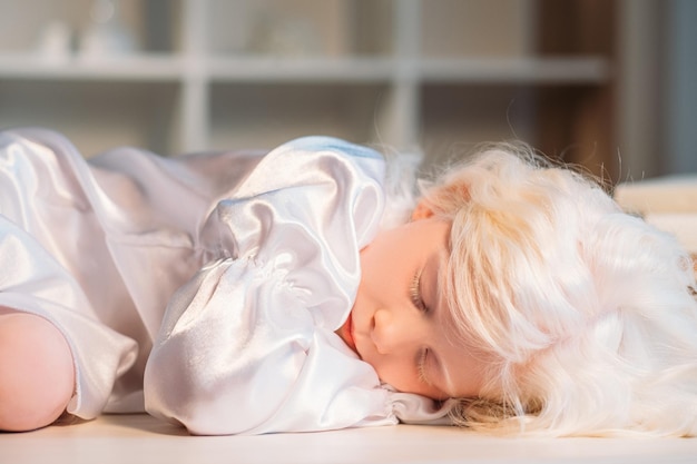 Siesta infantil Inocencia infantil Descanso tranquilidad Calma cansada linda rubia albina pequeña niña en vestido de noche de seda blanca acostada durmiendo con los ojos cerrados en el suelo sobre fondo de casa borroso