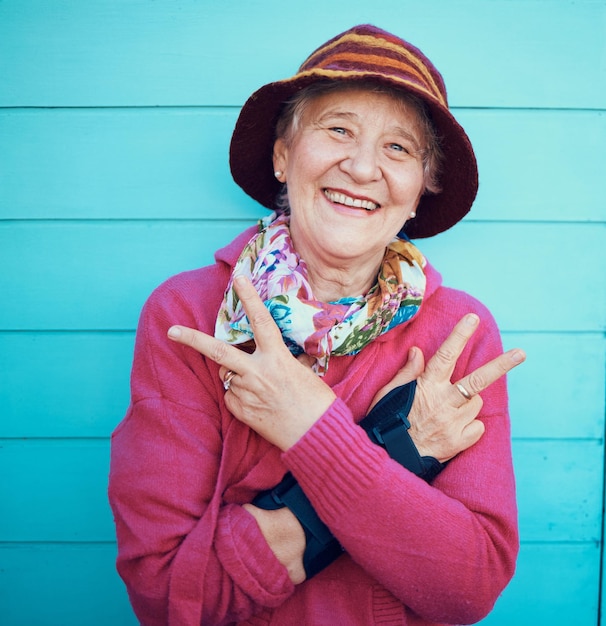 Signo de la paz hip hop y retrato de una anciana en la ciudad con sonrisa de energía y moda urbana Abuela de moda y anciana con emoji mano jubilada en un fondo azul o pared