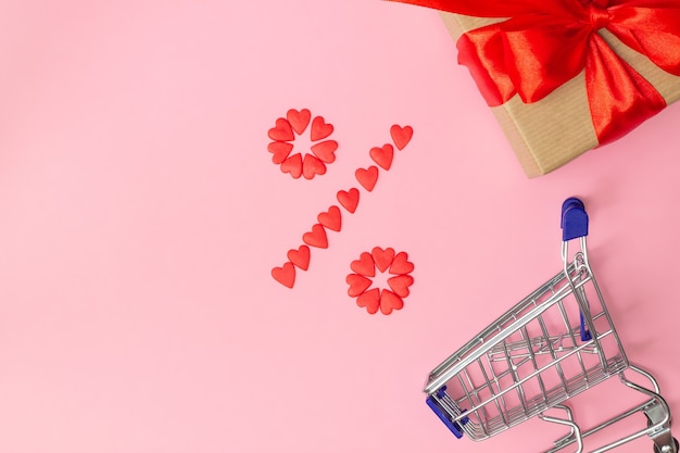 Signo de porcentaje forrado con pequeños corazones rojos con regalo envuelto en papel artesanal y cinta roja y carrito de compras sobre fondo rosa. Endecha plana. Copie el espacio.