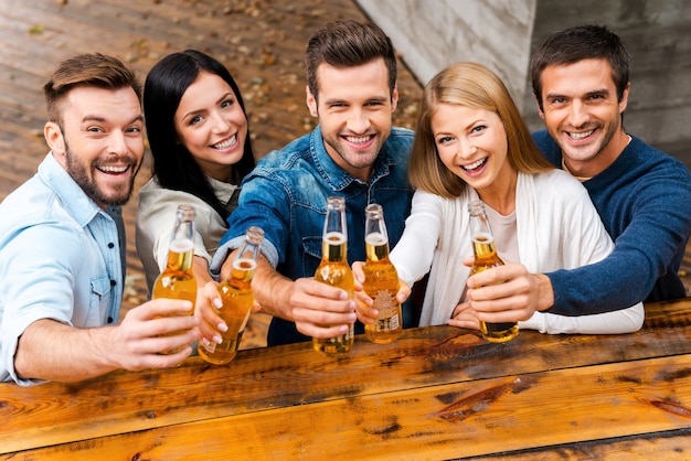 ¡Sigue la fiesta! Vista superior del grupo de jóvenes felices que se unen entre sí y estiran botellas con cerveza mientras están de pie al aire libre