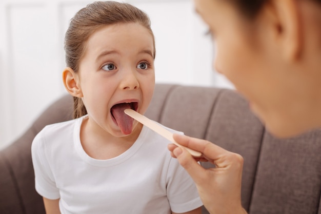 Siguiendo los comandos. Adorable y amigable niña haciendo lo que dice su médico y abriendo la boca mientras se somete a un chequeo médico