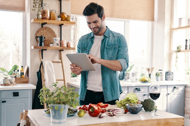 Siguiendo receta. Apuesto joven en ropa casual con tableta digital y sonriendo mientras está de pie en la cocina en casa