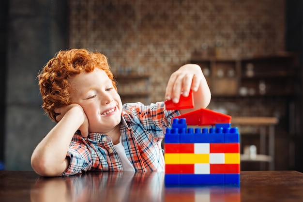 Siguiente gran arquitecto. Adorable niño descansando su atención en una mano y sonriendo ampliamente mientras está sentado en una mesa y disfruta jugando con un juego de construcción.