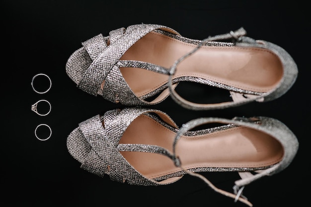 Silberne Schuhe und Ringe Zusammensetzung auf dem schwarzen Hintergrund isoliert Accessoire Braut Schuhe Trauringe und Sandale Schuh mit Kristall Nahaufnahme flach legen Ansicht von oben Verlobung