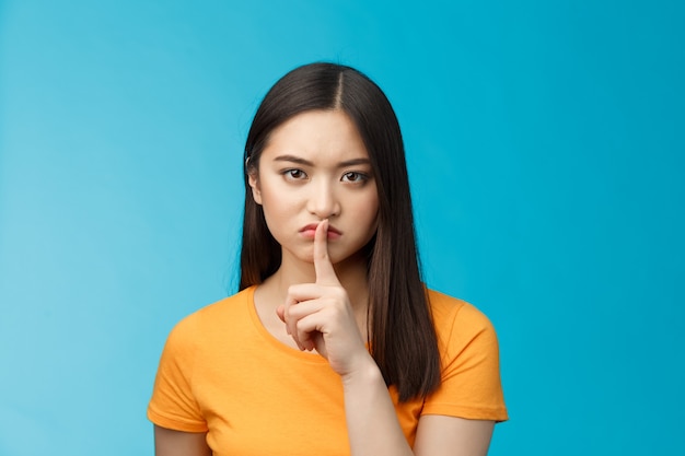 Silencio, quédate en silencio. Una chica asiática de aspecto serio se ve disgustada, gruñona, muestra el signo de silencio, presiona los labios del dedo índice, frunce el ceño molesto, dice que no habla, no dice secretos, prohíbe la música alta, fondo azul.