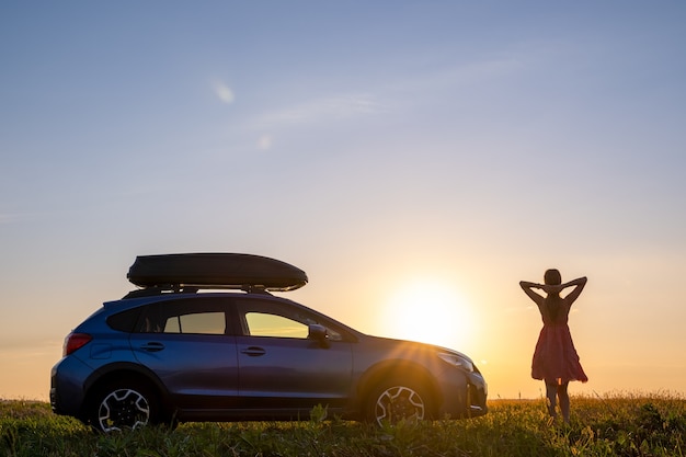 Silhouette der Fahrerin, die in der Nähe ihres Autos auf einer Wiese steht und den Blick auf den hellen Sonnenuntergang genießt. Junge Frau, die sich während der Fahrt neben dem SUV-Fahrzeug entspannt.