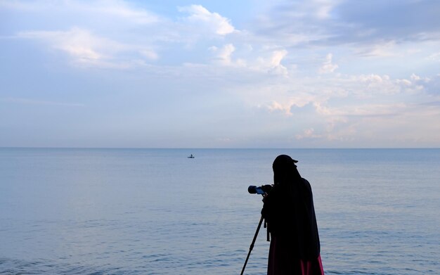 Silhouette der Frau im Hijab, die am Strand steht und den Horizont betrachtet