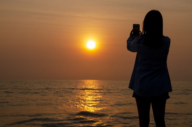 Silhouette der Frau machen Foto-Sonnenuntergang auf dem Meer und bunten Himmel mit dem Smartphone.