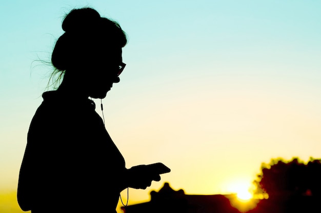 Silhouette der Frau mit Handy und Kopfhörern auf dem Hintergrund des Sonnenuntergangs. Social Media und Natur