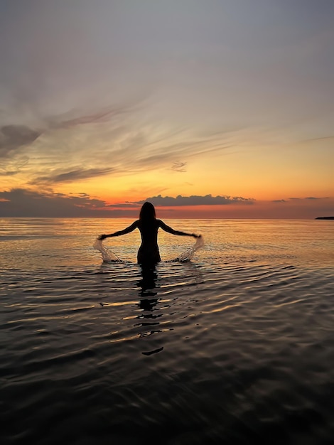 Silhouette der jungen Frau bei orangefarbenem Sonnenuntergang in der Meerwasserwelle und rosa bewölktem Himmel