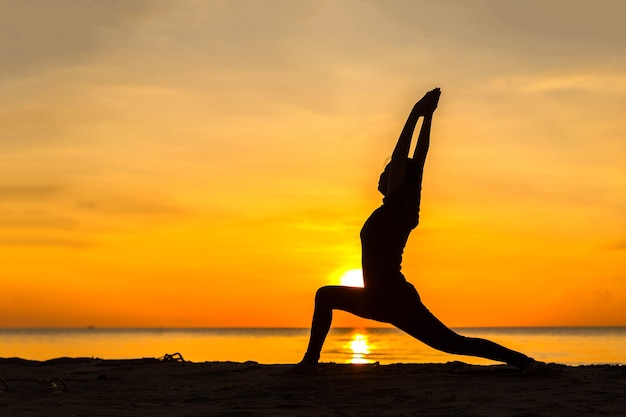 Silhouette der jungen Frau, die bei Sonnenuntergang Yoga am Strand praktiziert.