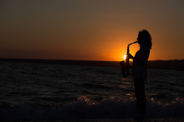 Silhouette der jungen sexy Frau, die bei Sonnenuntergang Saxophon am Strand spielt