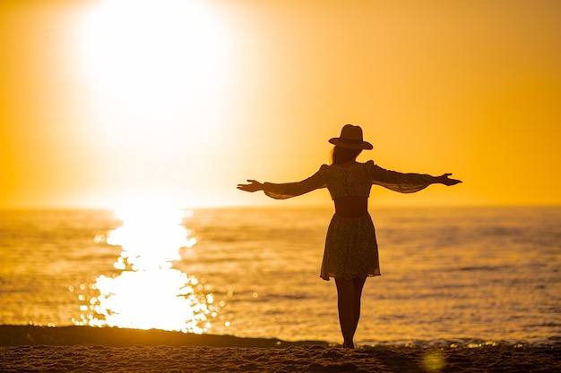 Silhouette der schönen Frau mit Strohhut, die einen wunderschönen Sonnenuntergang am Strand genießt