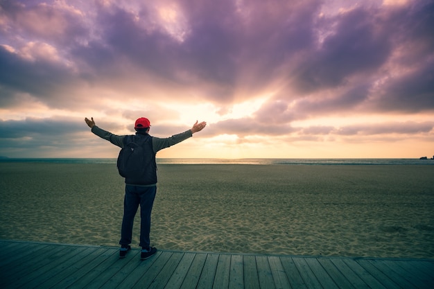 Silhouette des Mannes am Strand mit Blick auf den magischen dramatischen Sonnenuntergang. Mann mit Händen in der Luft, der auf der Holzterrasse steht