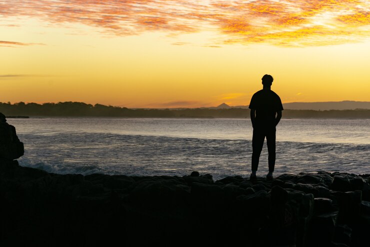 Silhouette des Mannes, der auf Felsen steht und den Sonnenuntergang mit einem Meerblick und einem Berghintergrund beobachtet.