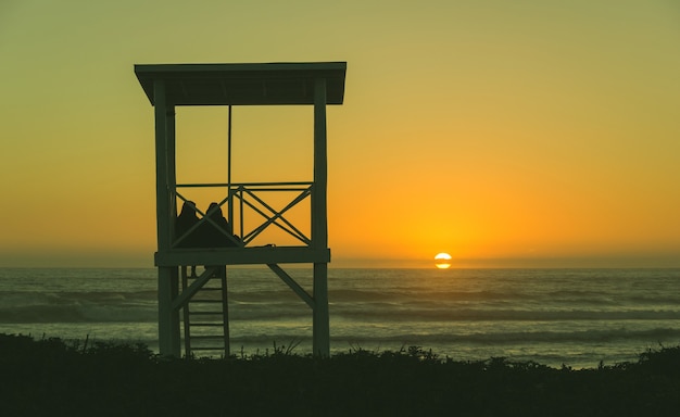 Silhouette des Rettungsschwimmerturms mit einem darauf sitzenden Paar beim Betrachten des Sonnenuntergangs in La Serena