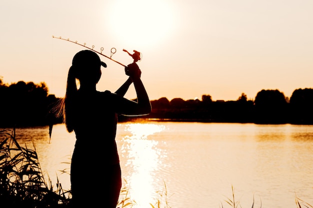 Silhouette einer Frau bei Sonnenuntergang mit einer Angelrute in der Nähe des Teiches