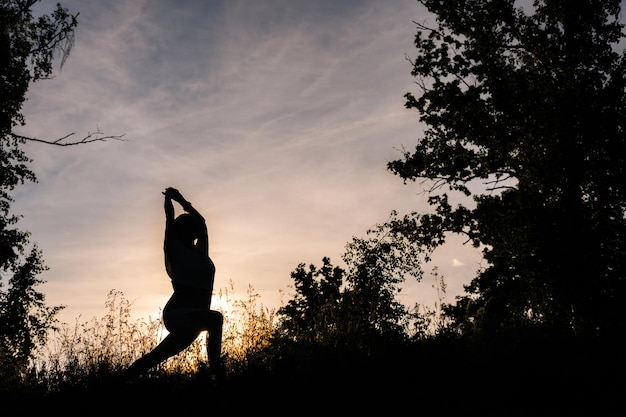 Silhouette einer Frau, die abends bei Sonnenuntergang Yoga-Asana-Pose eines Kriegers praktiziert
