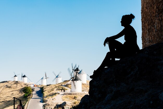 Silhouette einer Frau, die auf den Wänden einer mittelalterlichen Burg sitzt und antike Windmühlen in Toledo, Spanien, betrachtet.