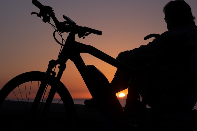 Silhouette einer Frau, die den Sonnenuntergang betrachtet, der mit dem Fahrrad am Meer sitzt und die Schönheit der Natur genießt